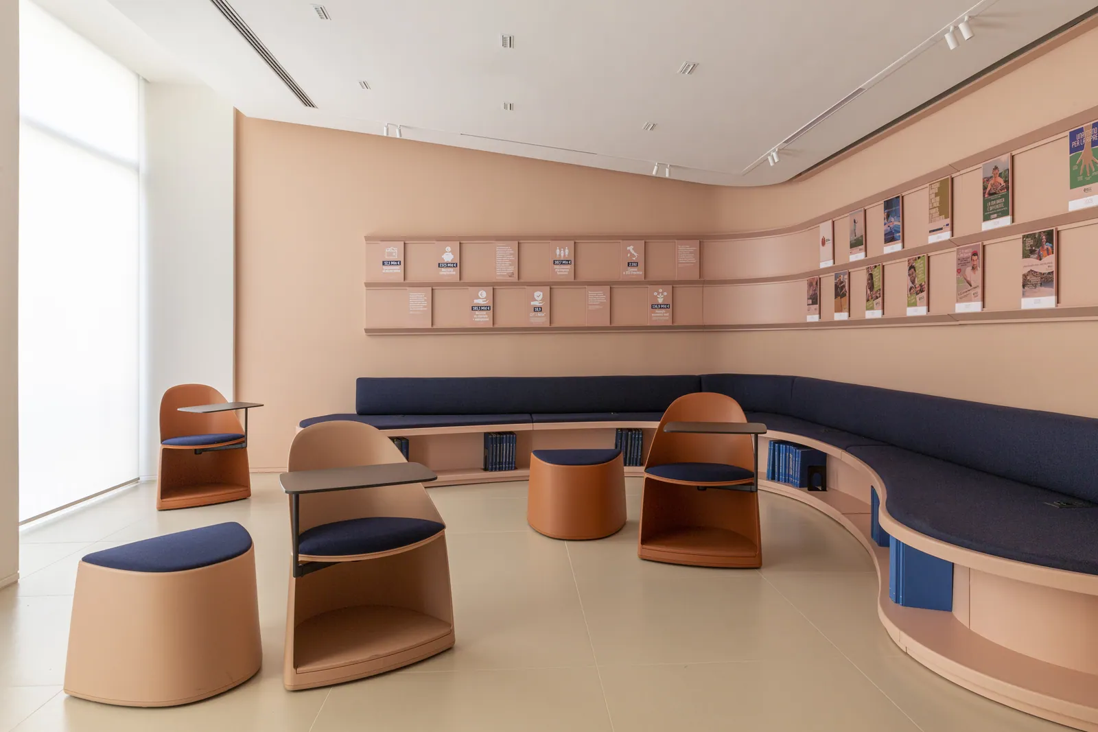 Sala con seduta continua curva blu, poltroncine e parete espositiva in tonalità cipria