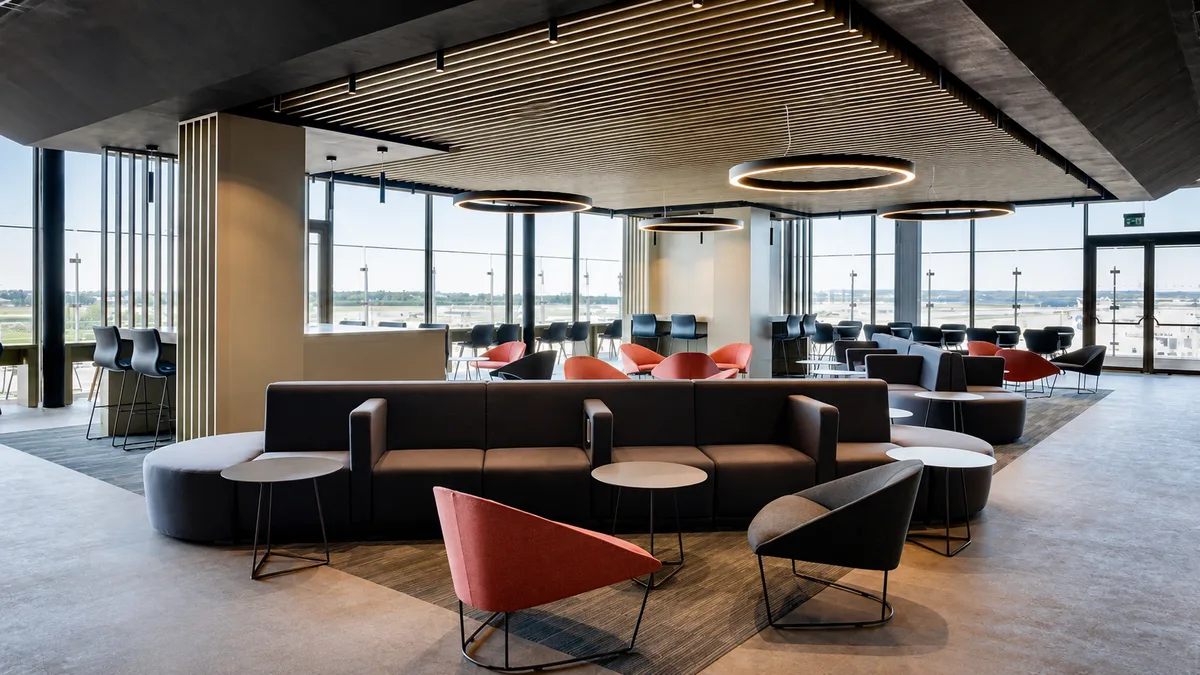 Area lounge con divani grigi, poltroncina arancio e vetrate a tutta altezza verso l'aeroporto