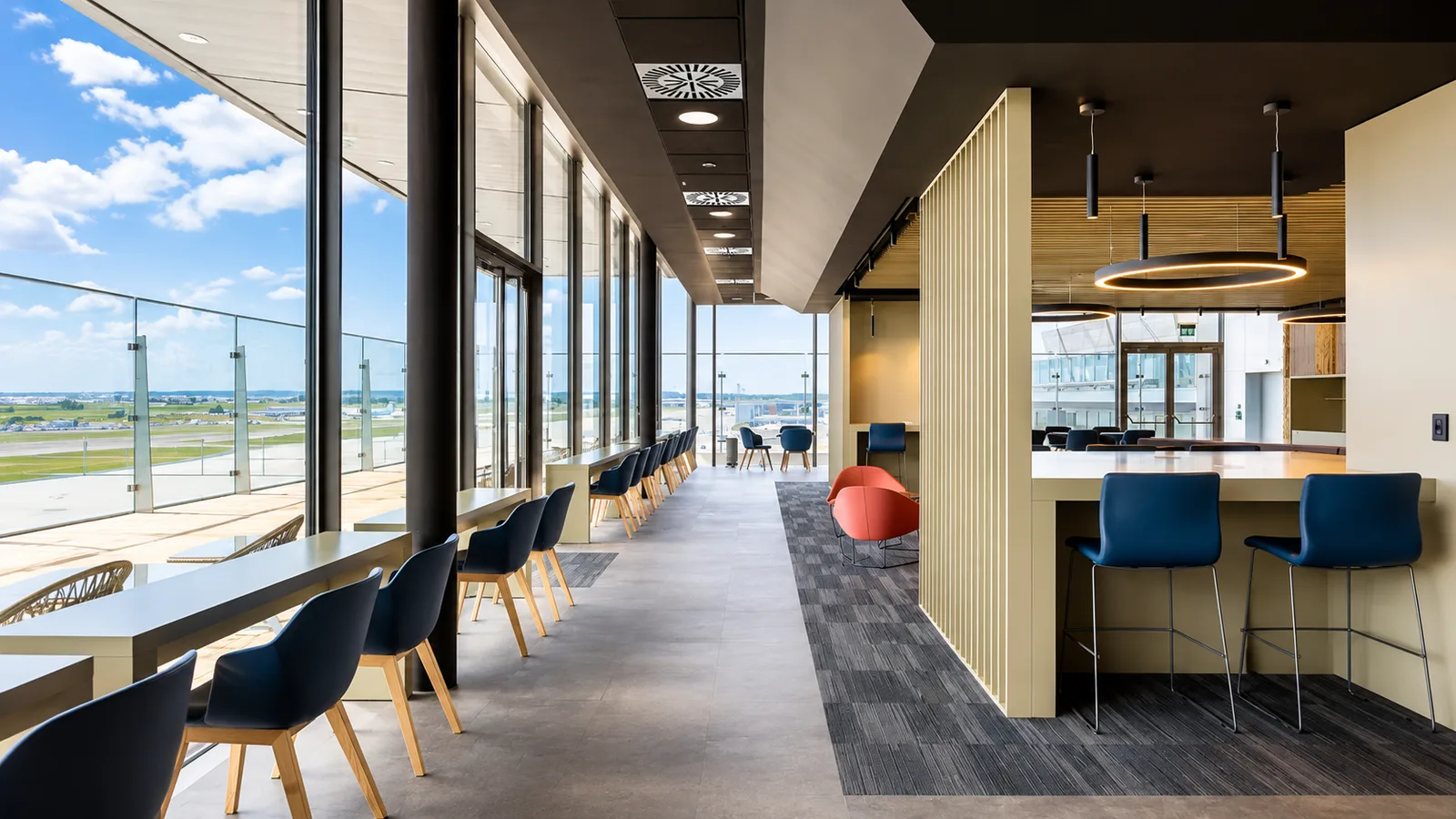 Sala d'attesa aeroportuale con bancone alto, sgabelli blu e vetrate affacciate sulle piste