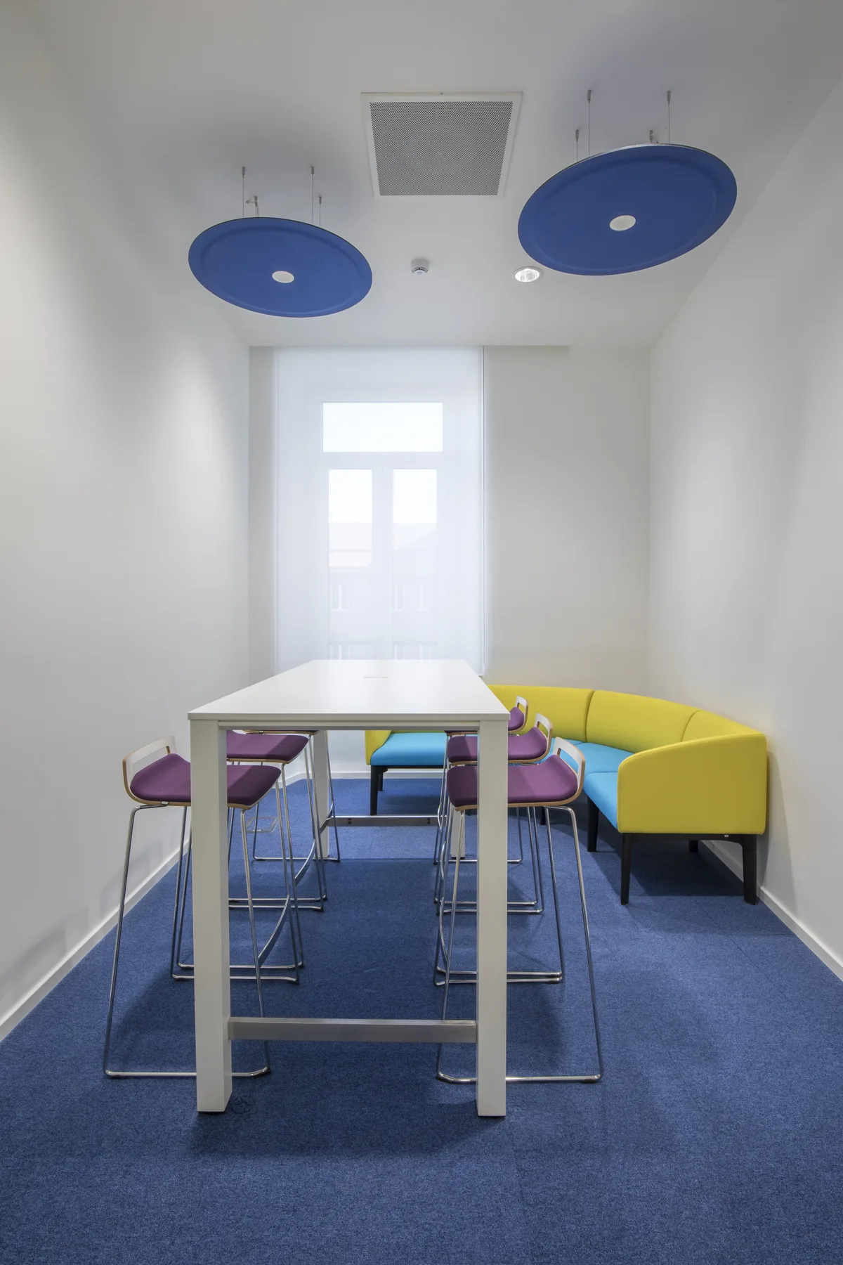 Area di lavoro informale con tavolo alto, sgabelli, panca gialla e moquette blu