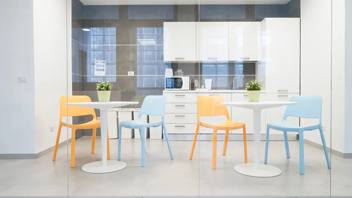Area cucina con mobili bianchi, tavoli rotondi e sedie arancio e azzurre