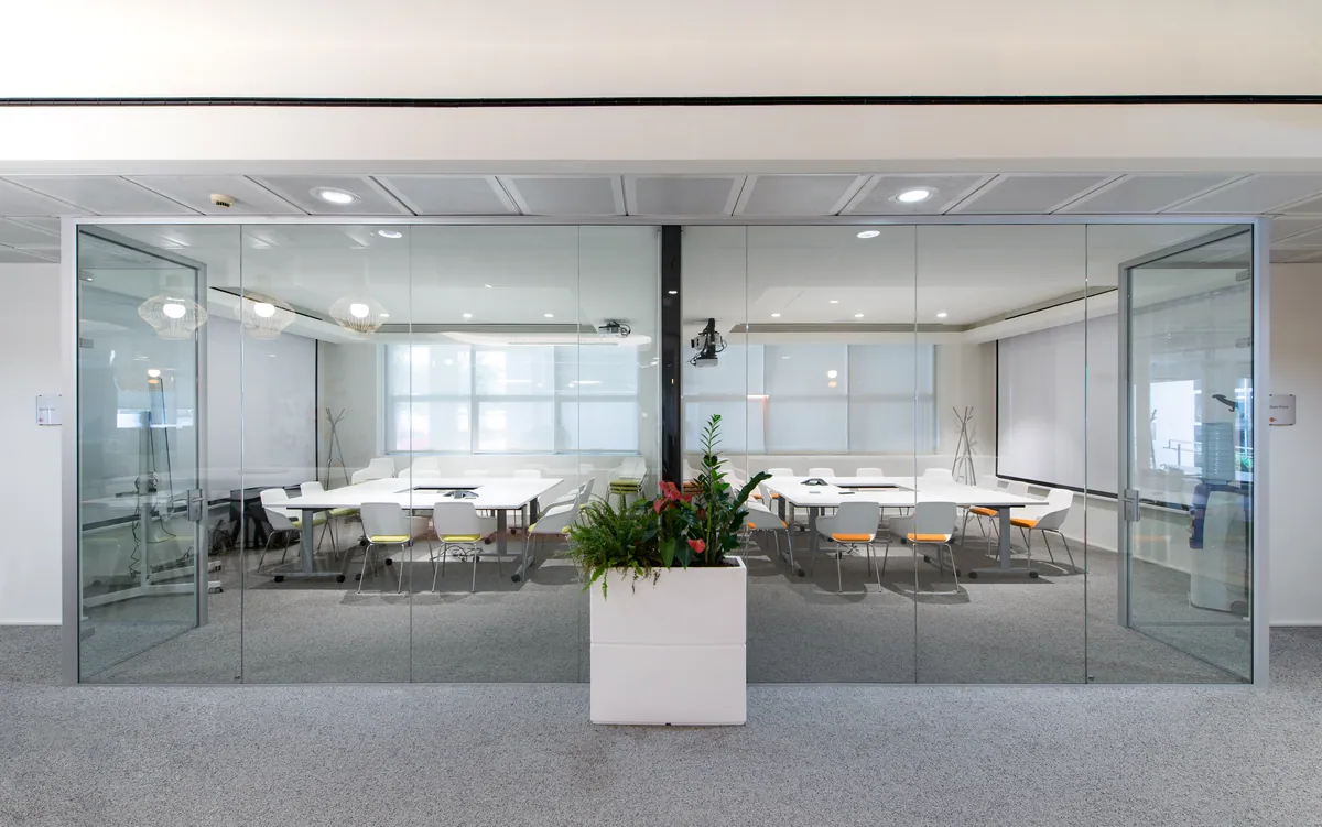 Sala formazione vetrata con tavoli modulari bianchi e fioriera centrale